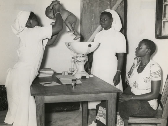 Village midwife, Rachel Yusuti (holding the baby), Mnyiramba and Ayah Brina Masasi, Msukuma, Weighing an infant and giving advice to the mother during an infant welfare clinic session at Kishapu Native Authority Dispensary and Clinic in Shinyanga District, Lake Province - Catalogue reference: CO 1069/157