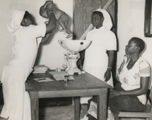 Village midwife, Rachel Yusuti (holding the baby), Mnyiramba and Ayah Brina Masasi, Msukuma, Weighing an infant and giving advice to the mother during an infant welfare clinic session at Kishapu Native Authority Dispensary and Clinic in Shinyanga District, Lake Province - Catalogue reference: CO 1069/157