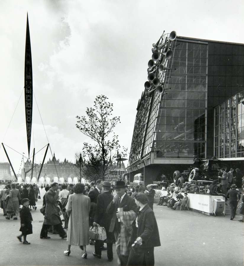 A tall, narrow, tapered structure – the Skylon – is held in place by wires next to a glass-fronted building of a similar height.
