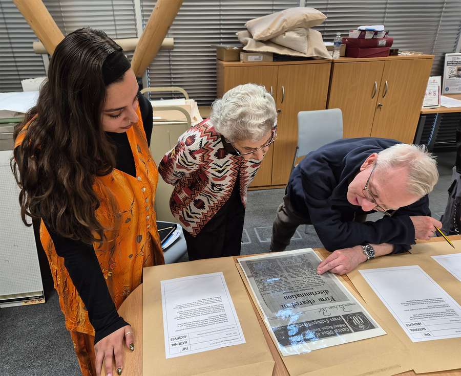 Three people gather round a laminated copy of a newspaper article, reading it closely.