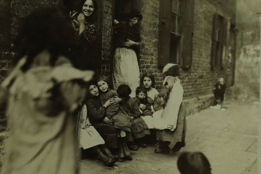 Photograph of a poor 19th-century family sitting on a doorstep in a London street. The children are smiling at the camera.