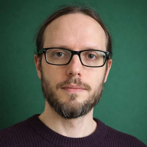 Andrew is looking towards the viewer. He has brown hair, a beard and glasses. He is also wearing a purple jumper and standing in front of a green background.