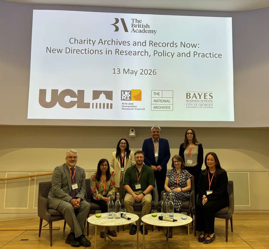 Photograph of eight people seated and standing in front of a large screen displaying logos of The British Academy, UCL, UKRI, The National Archives, and Bayes Business School.