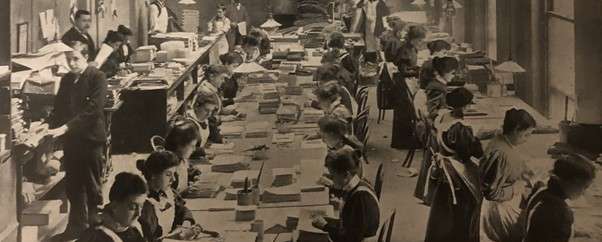 A large room full of men and women working at benches wearing white aprons