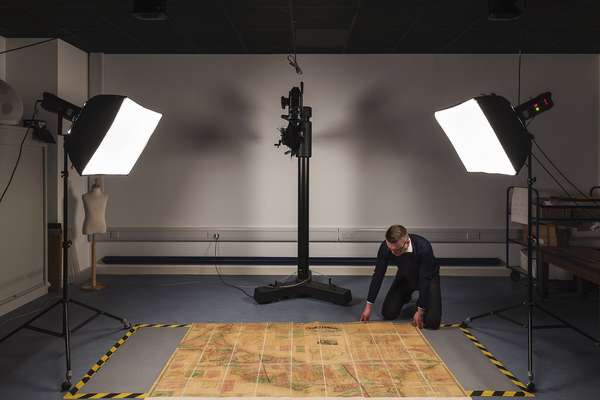 A person kneels on the floor adjusting a large historical document ready for it to be photographed. It is illuminated by two bright lights.