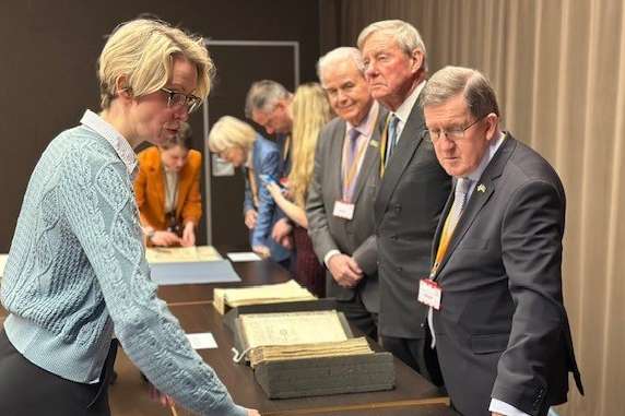 A group of dignitaries look in interest at a range of historical documents on a table.