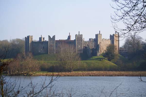 An impressive stone castle with five evenly-spaced towers, on top of a high grassy mound, beside a lake.