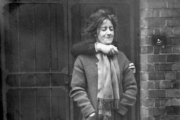 Photograph of a woman standing in a yard with a defiant look on her face. An unseen figure stands behind her with their arm around her neck restraining her from behind.