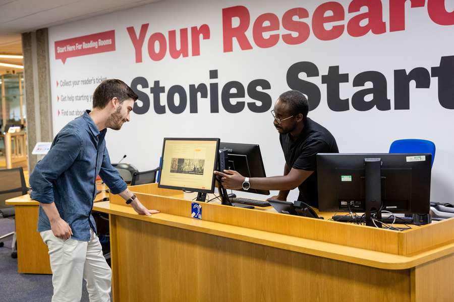 A visitor stands at a help desk and is shown information on a computer by a member of staff.