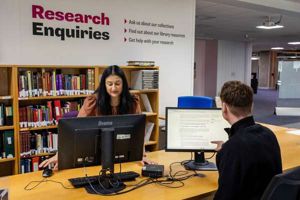A helpdesk with a sign saying 'Research enquiries'. A member of staff shows a visitor some helpful information on a computer screen.