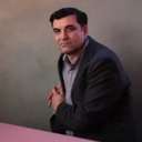 A man with dark hair in a suit is sat in a chair, facing the viewer.