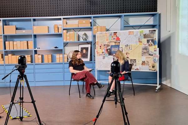 Two people sit in a broadcast studio in front of a board featuring cut out records and images..
