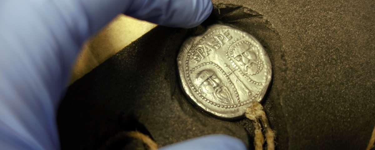 A blue-gloved hand reaches out to touch a metal seal encased in foam and depicting the faces of two men.