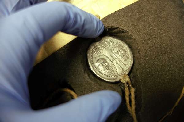 A blue-gloved hand reaches out to touch a metal seal encased in foam and depicting the faces of two men.