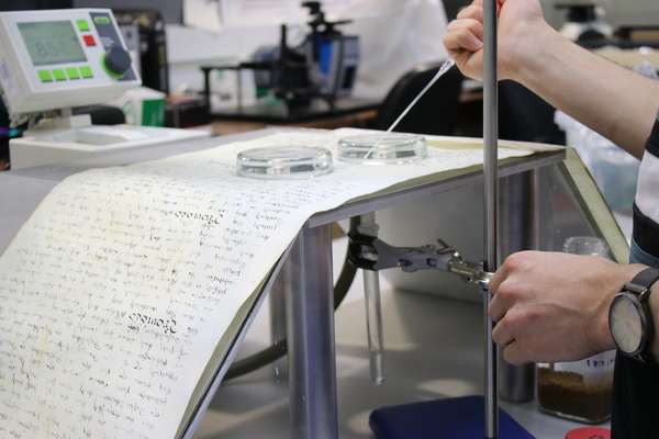 A parchment roll is laid suspended across a table while a scientist performs analysis.