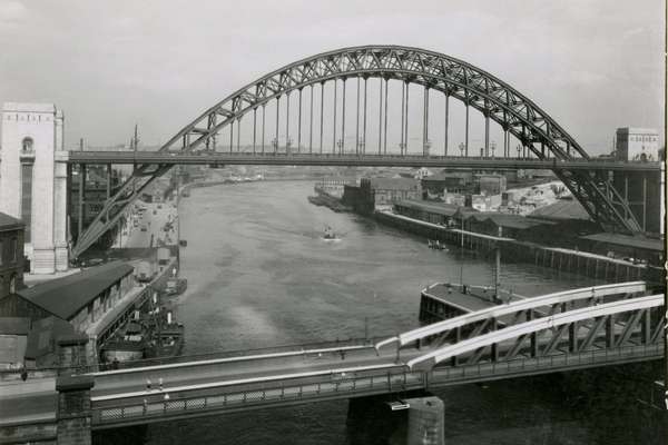 Photograph of the Tyne Bridge, a large through arch bridge positioned high over the river. A smaller bridge is visible in the foreground.