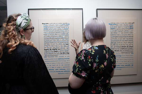 Two women stand in front of two large portrait embroideries with hand-stitched words in thread.
