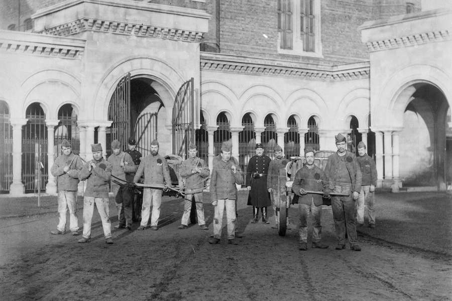 A group of prisoners stand in a line to be photographed. They're standing in a yard in front of an imposing building with tall walls and arches. The men are holding the handles of carts and wearing prison uniforms.