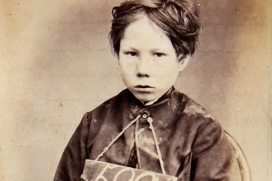 Photograph of a young boy with a prison number written on a board around his neck. He sits with his hands on his knees looks sullenly into the camera.
