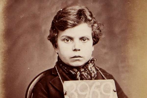 Photograph of a young boy sat with a prisoner number written on a board that hangs around his neck. He looks defiantly into the camera.