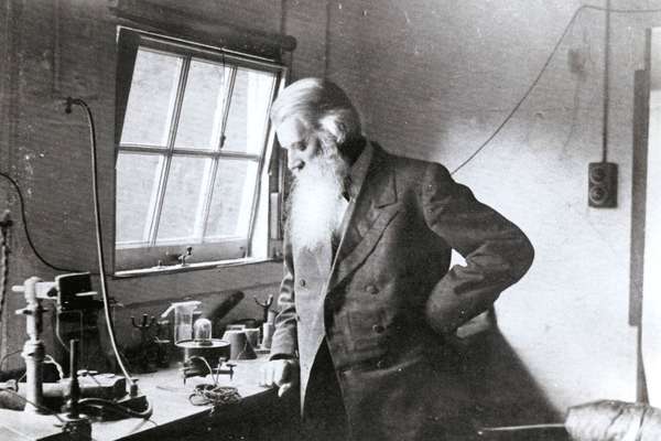 A Victorian man with a long white beard looks down at a desk filled with scientific equipment.