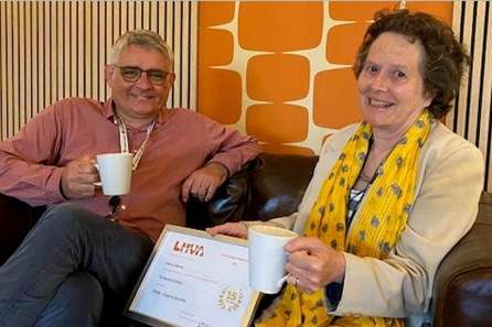 A smiling man and a woman sit on a sofa with cups of tea. Between them is a framed certificate.