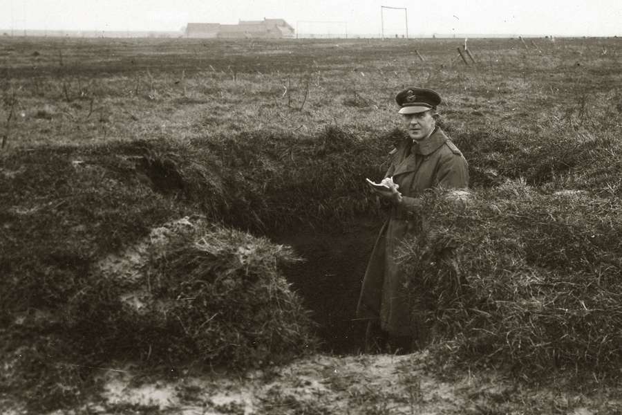 Photograph of a First World War soldier standing in a hold in an open field taking notes on a notepad.