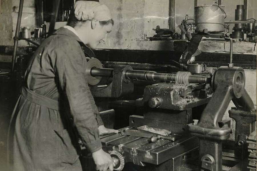 A woman with her hair held back in a scarf, uses heavy machinery in a factory.