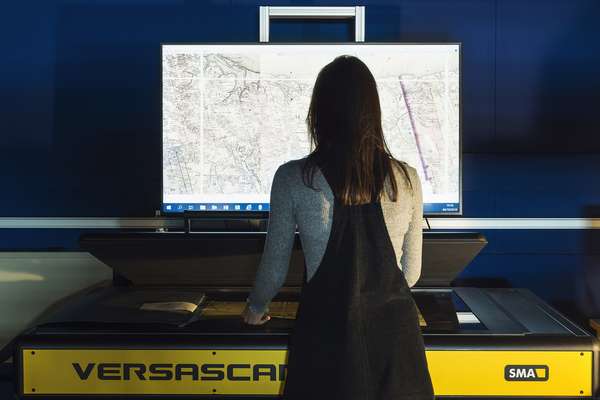 A person stands at a large industrial scanning machine. A historical document is shown on a computer screen above.