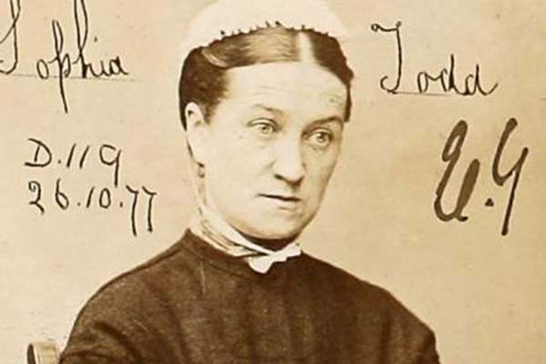 A woman in a black dress with white cap sits with her arms folded in her lap