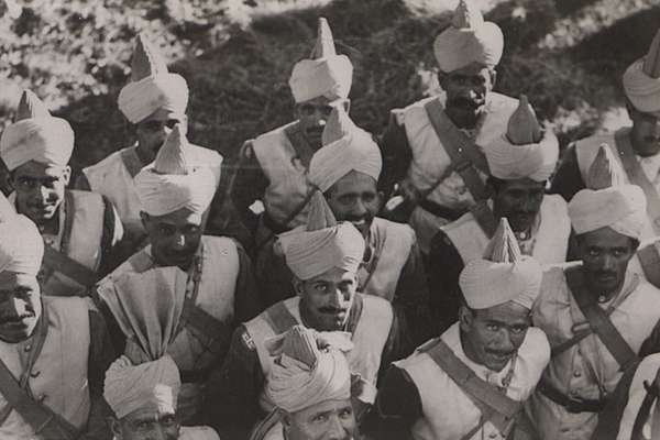 Monochrome photo of a group of South Asian men dressed in uniform. Many look up at the camera and some smile.