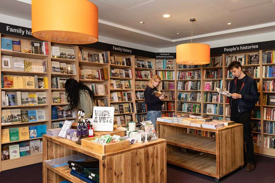 Customers browse books and gifts in a shop. The walls are lined with bookshelves with category labels such as Family History and People's History.