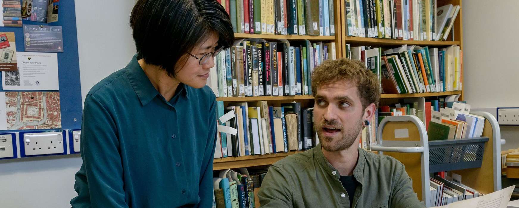 A man in a green shirt is sitting at a table, talking to a woman in a teal shirt, who is standing next to him. Several documents and a laptop are on the table.