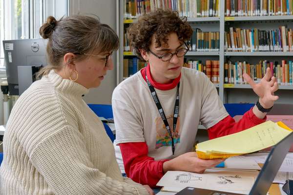 Two people sitting at a desk, sharing a laptop. They are both looking at a yellow document that the person on the right is holding.