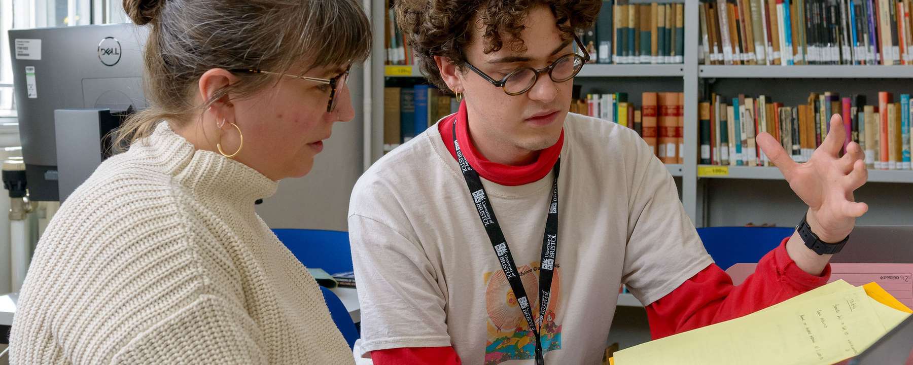 Two people sitting at a desk, sharing a laptop. They are both looking at a yellow document that the person on the right is holding.