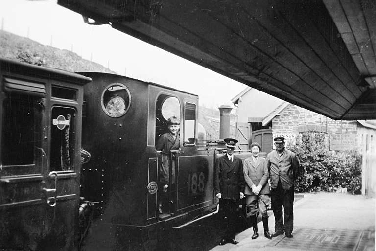 Photograph of a group of people standing next to a steam train at a station.