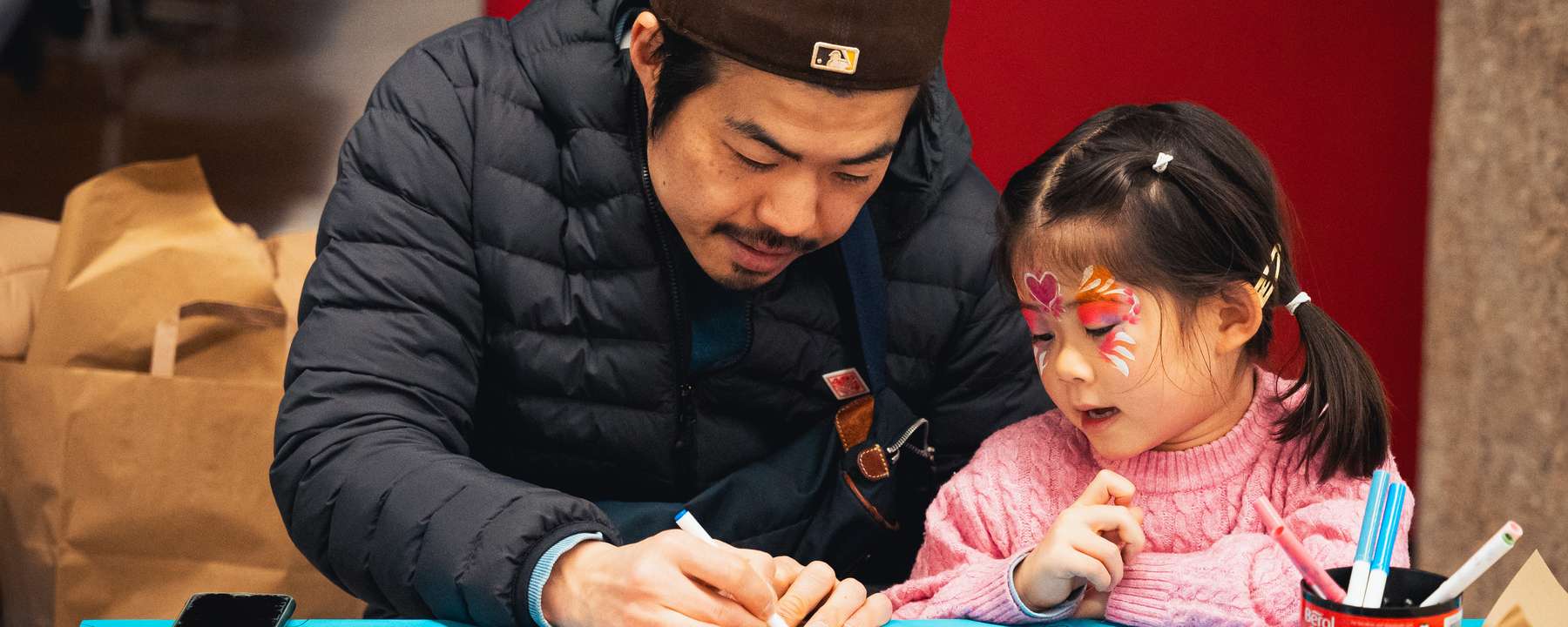An adult sketches on a piece of card while a child with their face painted watches intently.