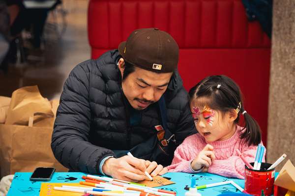 An adult sketches on a piece of card while a child with their face painted watches intently.