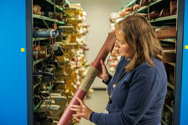 A member of staff holds a wrapped tube with both hands, in front of archive shelving packed with similar documents in tubes.