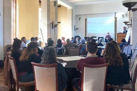 Groups of 8 sit around tables and listen to someone speaking in front of a projector.