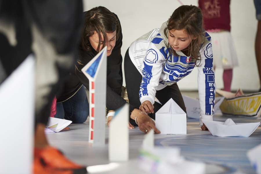 An adult and a child place a small, folded paper house on a mat near other paper buildings and a paper boat.