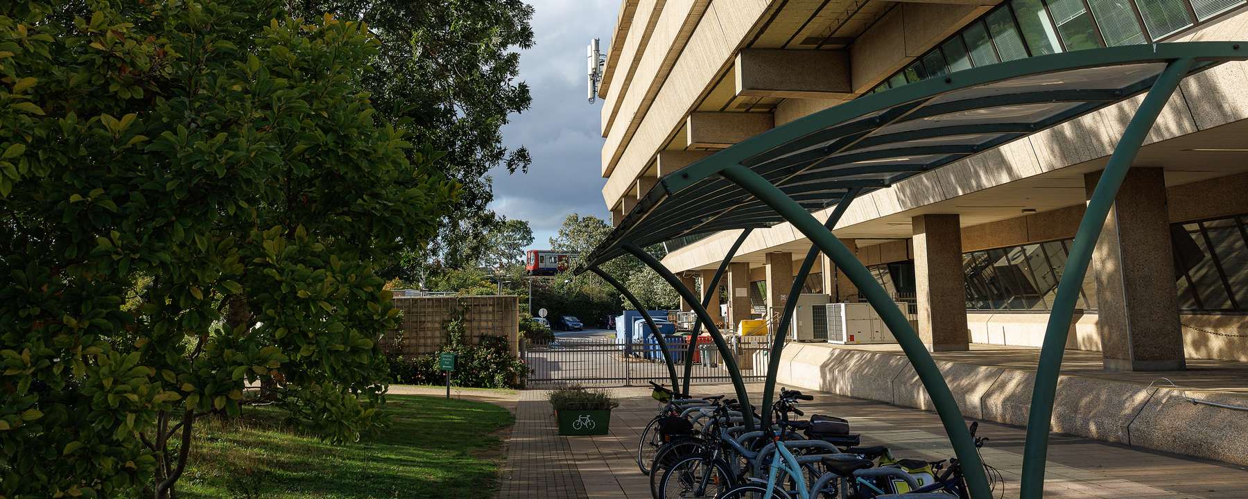A covered bike shelter sits between The National Archives Building and a grassy bank.