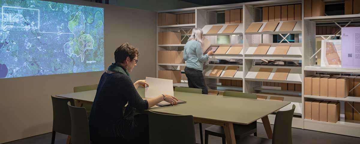 Two people look through files of documents in a space surrounded by shelves full of books and files