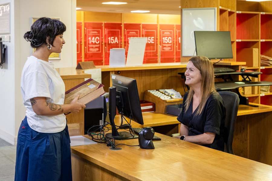 A visitor stands in front of a smiling member of staff who is sat behind a wooden desk with a computer screen on it.