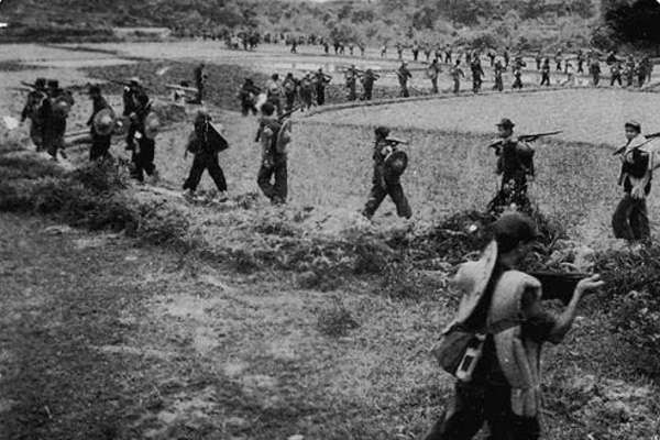 A photograph of a military group marching one by one in a line through fields.