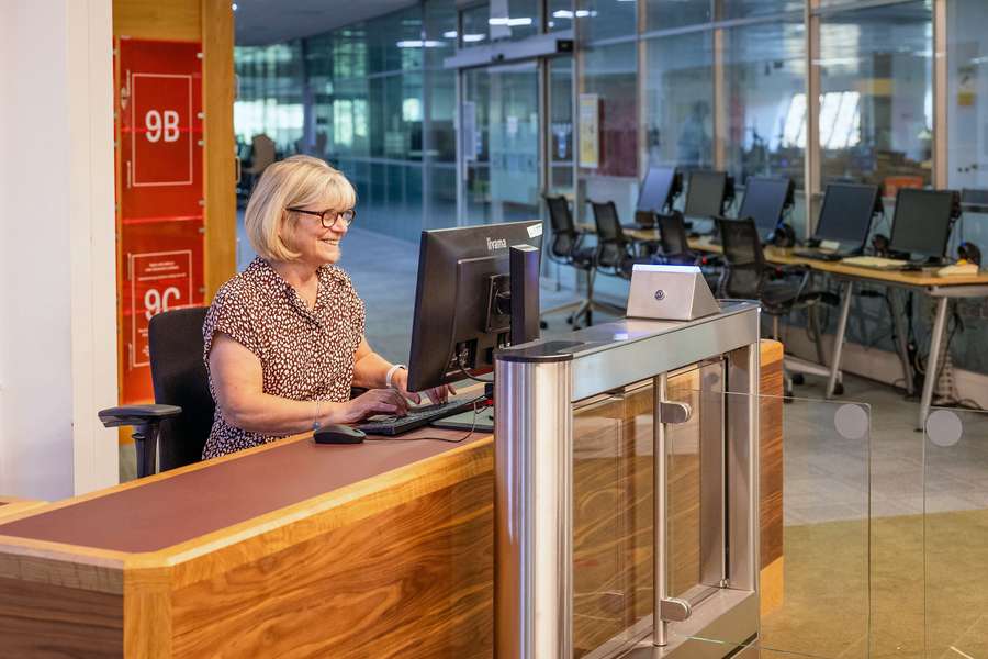 A smiling member of staff sits behind a desk using a computer, in front of an automatic glass barrier attached to a card scanner.