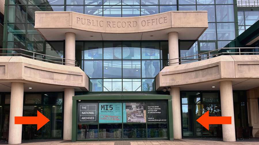 Front entrance of The National Archives. The building has large glass windows and concrete pillars. A sign above the entrance reads 'PUBLIC RECORD OFFICE'.