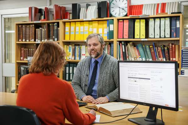 visitor sits down opposite a member of staff next to a computer screen and an open historical volume.