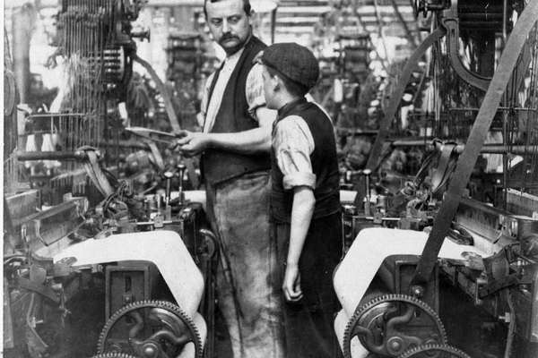 A man in a waistcoat looks down at a boy wearing a flat cap in the aisle between mill equipment.