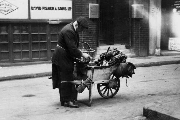 A man wearing a flat cap and holding a basket reaches into the wheelbarrow in front of him.